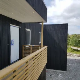 Nowoczesny dom z czarną, drewnianą elewacją, jasną drewnianą balustradą balkonu i zabezpieczonymi folią drzwiami wejściowymi, widok na krajobraz w tle.