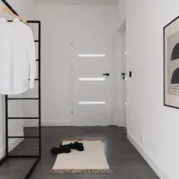 Minimalistyczny przedpokój z białą, nowoczesną drzwiami, czarnym wieszakiem na ubrania z wiszącą białą koszulą, geometrycznym obrazem na ścianie, ciemną podłogą i jasnym dywanikiem z frędzlami.