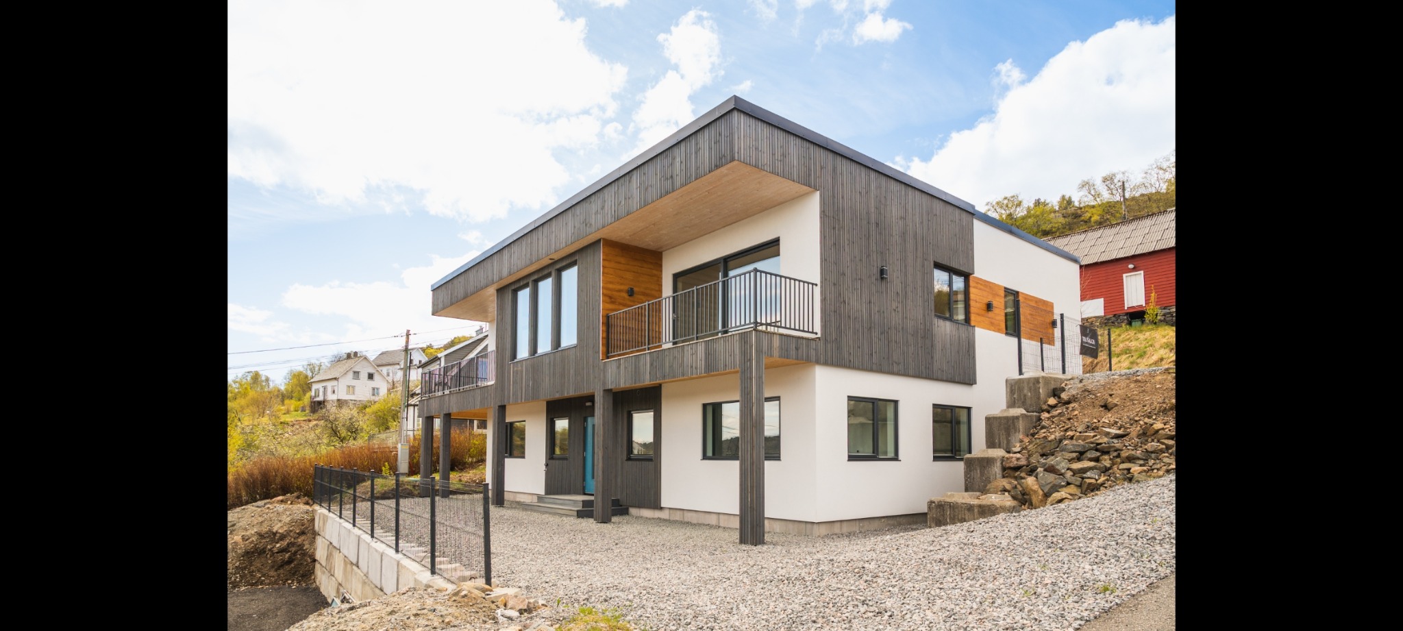 Nowoczesny dwupiętrowy dom z elementami drewnianymi, białymi ścianami i balkonem z czarną balustradą, otoczony żwirowym podłożem i niskim ogrodzeniem.