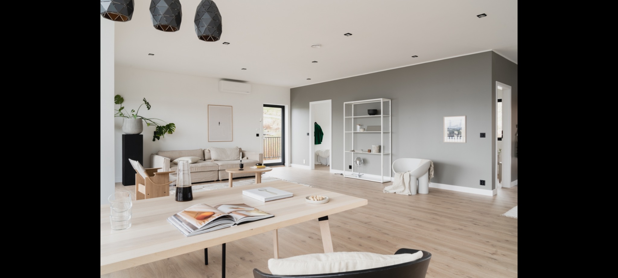 Jasne, przestronne wnętrze domu z drewnianą podłogą, beżową sofą, minimalistyczną półką i stołem z otwartą książką kucharską.