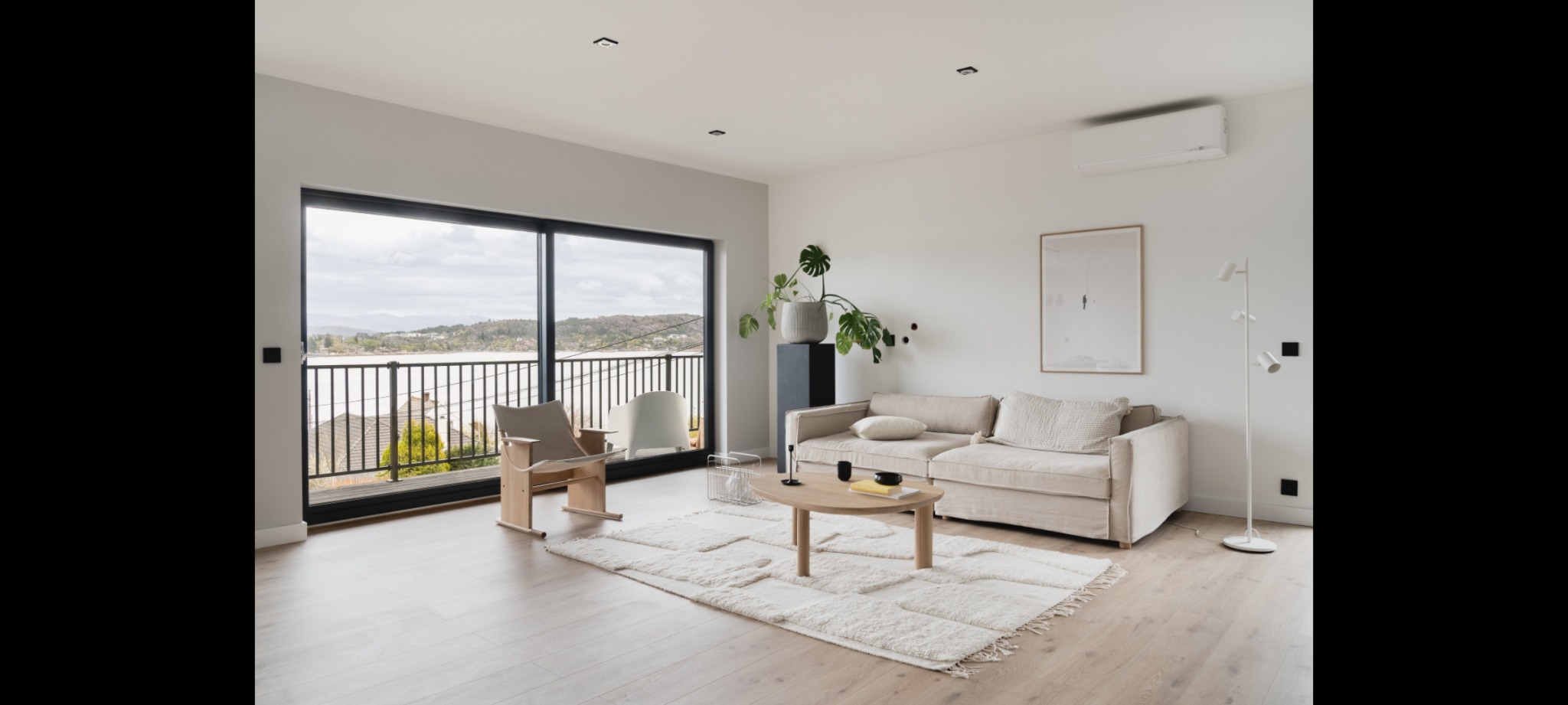 Jasne wnętrze salonu z dużym oknem balkonowym z czarną ramą i widokiem na jezioro z balustradą, beżowa sofa, drewniany stolik kawowy na puszystym dywanie, fotel z jasnego drewna i roślina w doniczce.