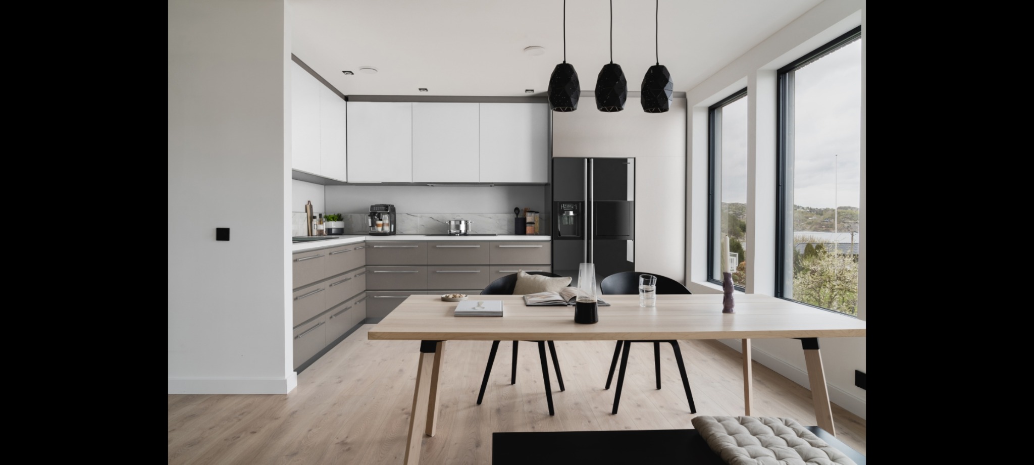Jasna, nowoczesna kuchnia z drewnianym stołem, czarnymi krzesłami i lampami wiszącymi o geometrycznym kształcie. Widok na zewnątrz przez duże okna.