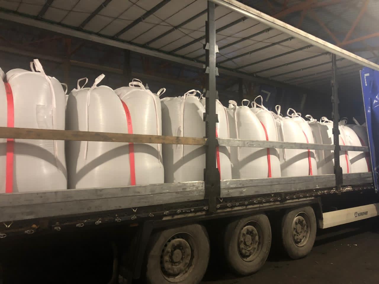 Ciężarówka z naczepą załadowana rzędem dużych, białych worków z czerwonymi pasami, zabezpieczonych drewnianą belką, widziana z boku w słabym oświetleniu magazynu.