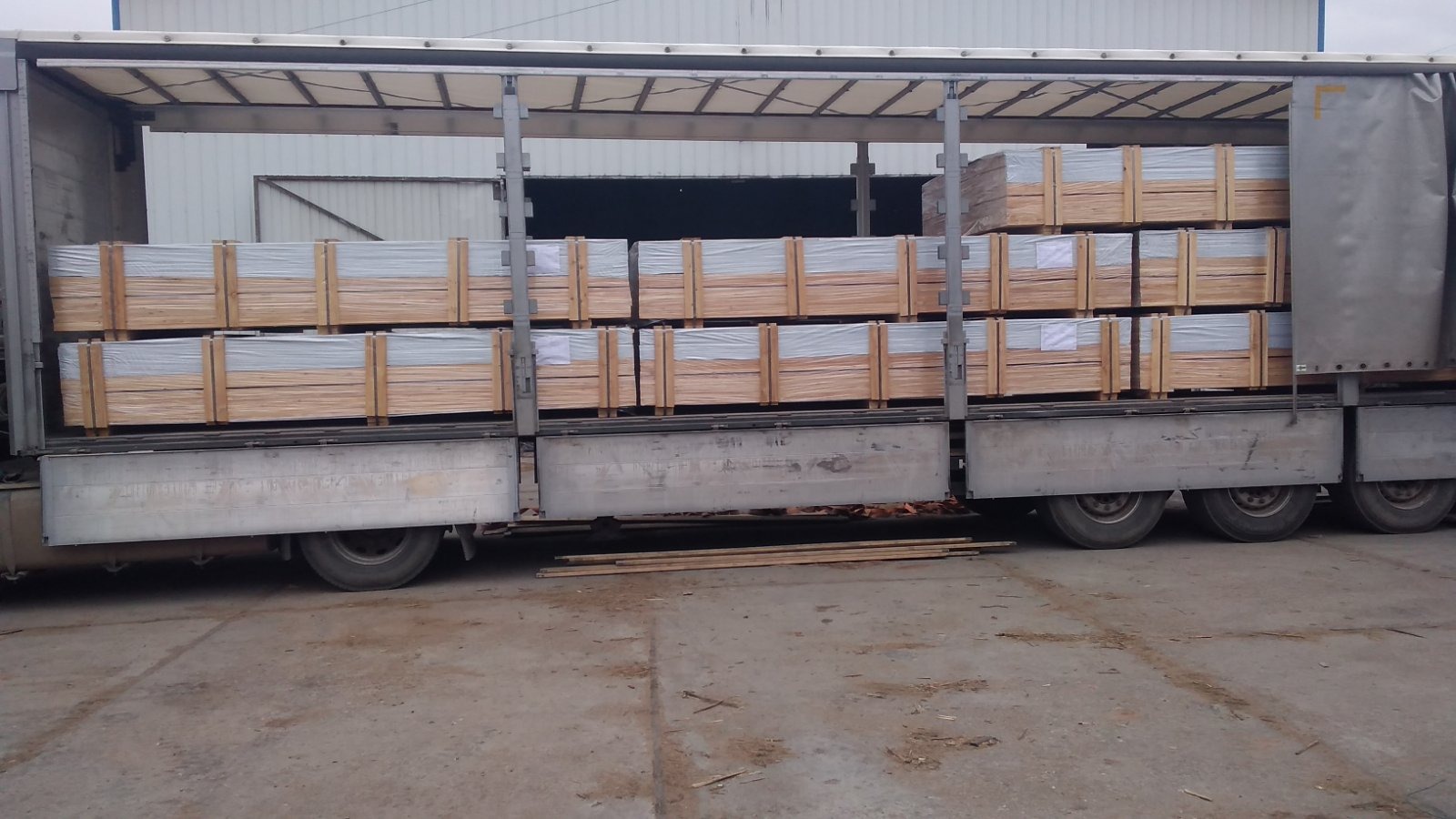 Ciężarówka z otwartą naczepą, załadowana paletami z drewnianymi skrzyniami, zabezpieczonymi folią, widok z boku na betonowy plac.