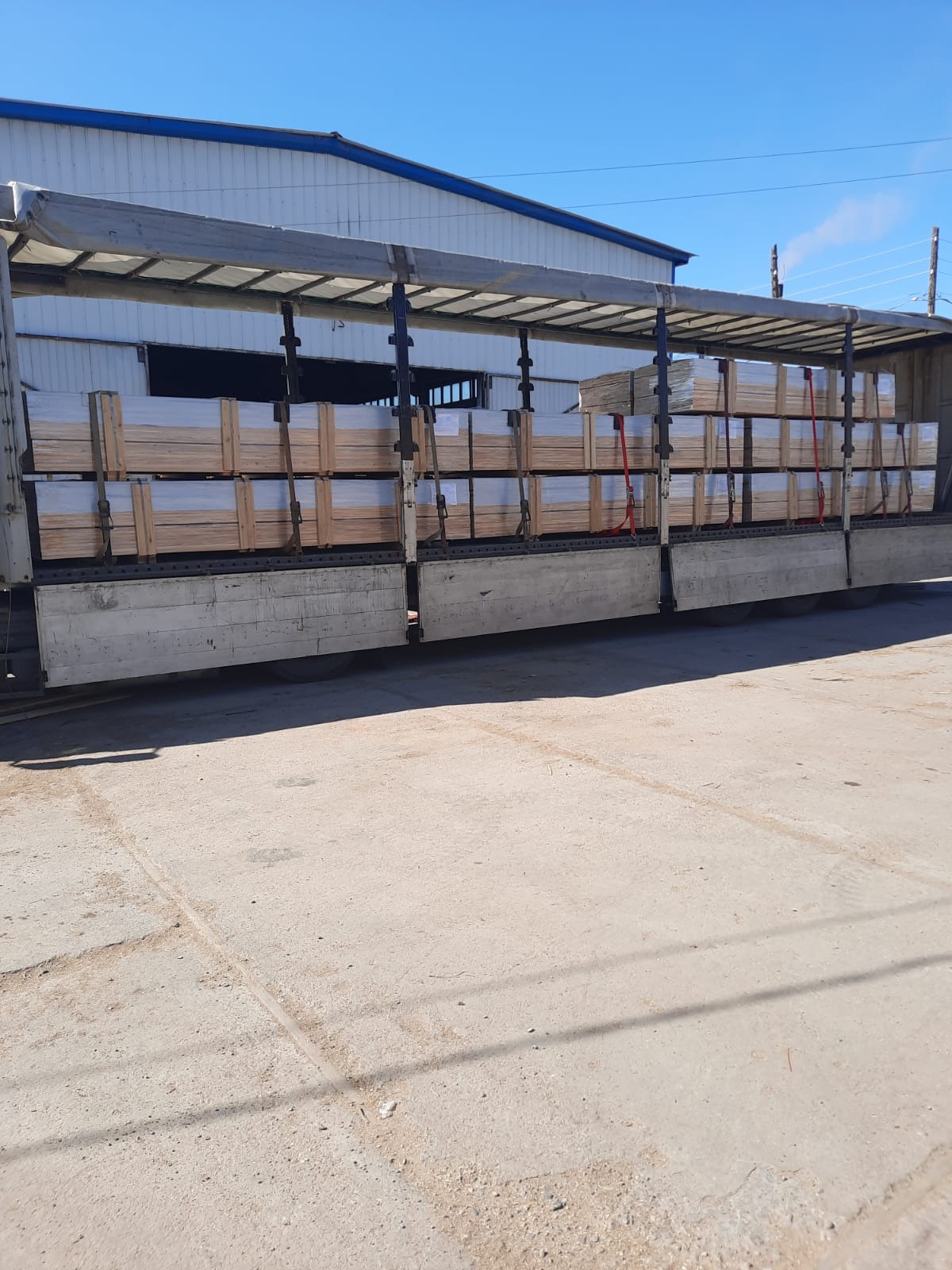 Ciężarówka z otwartą naczepą załadowana paletami z drewnem, zabezpieczonymi czerwonymi pasami transportowymi, na tle budynku magazynowego z niebieskim dachem.