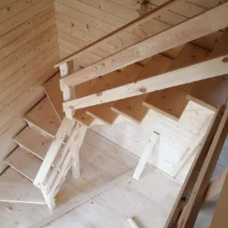 Wnętrze domu w trakcie budowy, ukazujące drewniane schody z surowego drewna, zamontowane w pomieszczeniu wykończonym boazerią, widoczne fragmenty podłogi i ścian z desek.