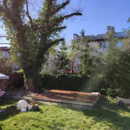 Podkonstrukcja pod przyszły taras z desek kompozytowych, widoczne narzędzia malarskie i puszka farby, trawa i duży drzewo w tle.