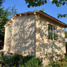 Nowo wybudowany, jasny, drewniany domek letniskowy z oknem i widocznymi złączeniami bali, otoczony zielenią i częściowo zacieniony przez liście drzew.