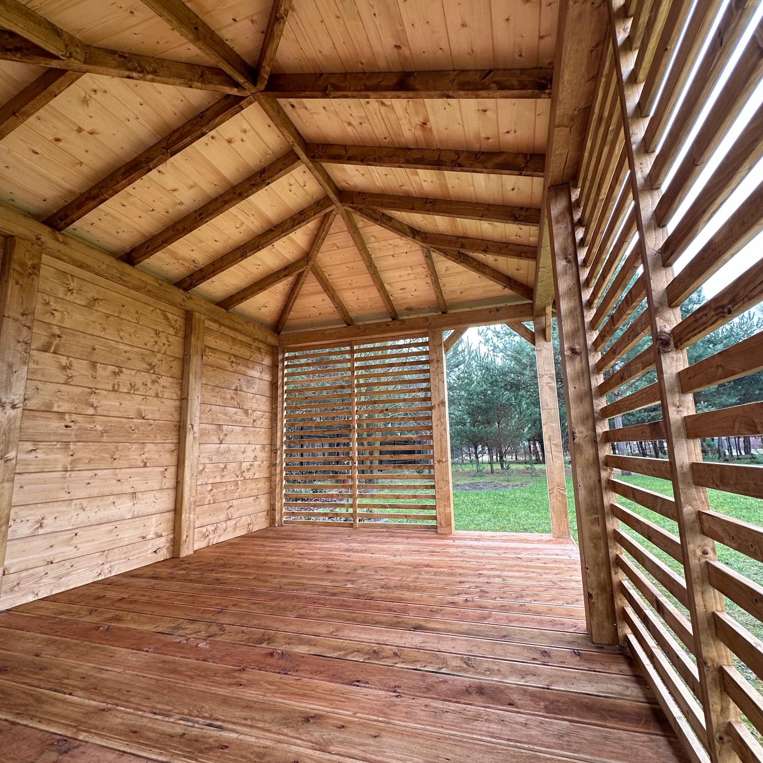 Drewniana konstrukcja z ażurowymi ścianami, widok z wnętrza, ukazujący podłogę z desek i skośny dach, zieleń w tle.