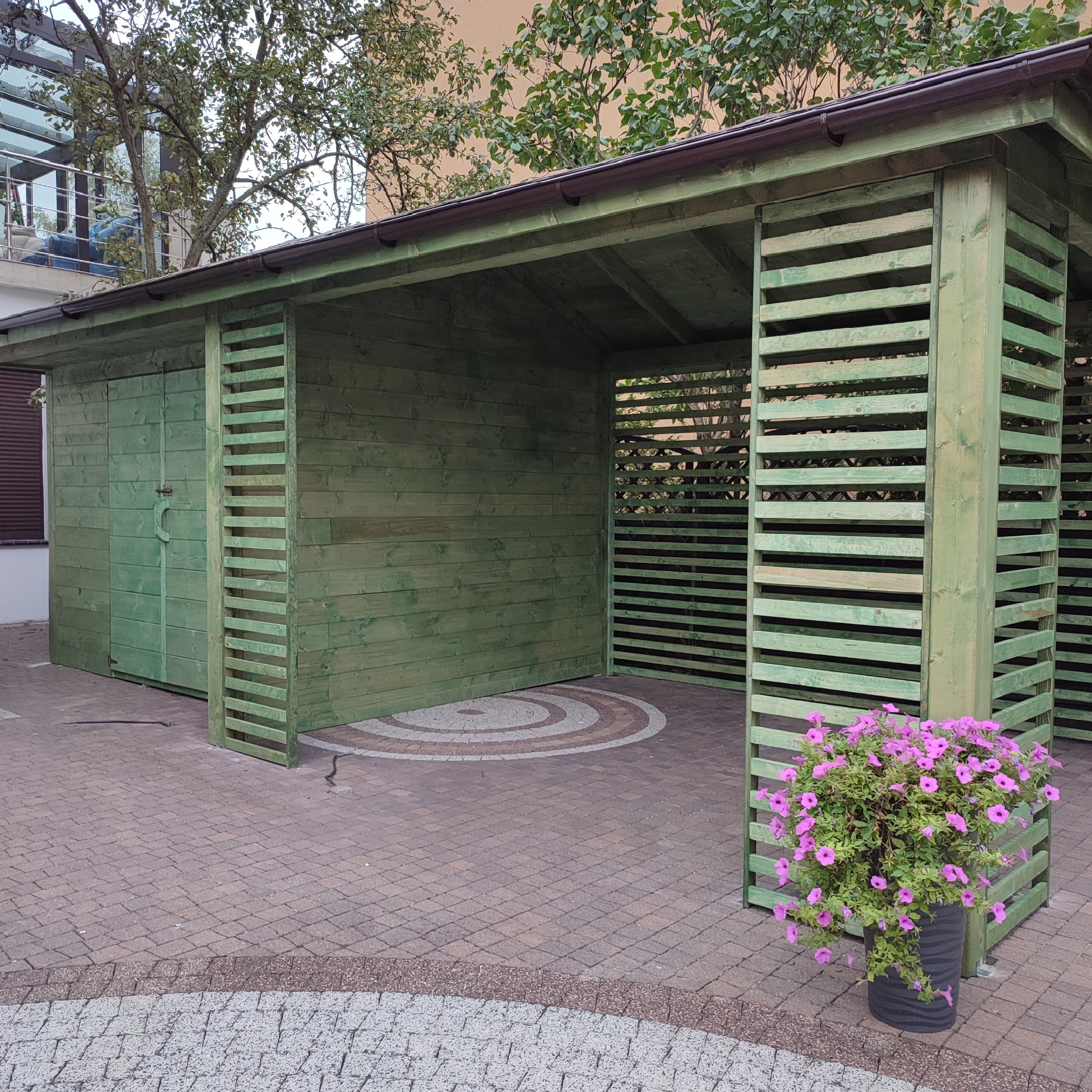 Zielona, drewniana konstrukcja z zamykaną częścią i otwartą przestrzenią z ażurowymi ścianami, stojąca na brukowanym podłożu, z donicą z fioletowymi kwiatami z przodu.