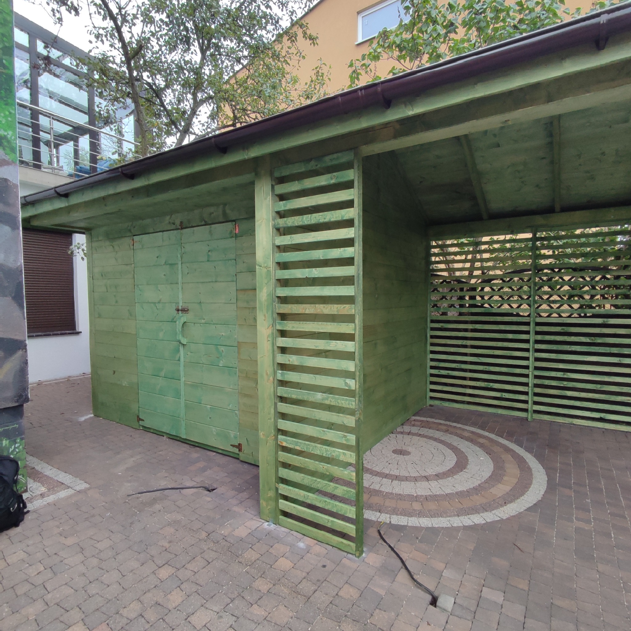 Zielona drewniana konstrukcja z zamykanymi drzwiami i ażurowymi ścianami, zlokalizowana na brukowanym podłożu z dekoracyjnym wzorem koła.