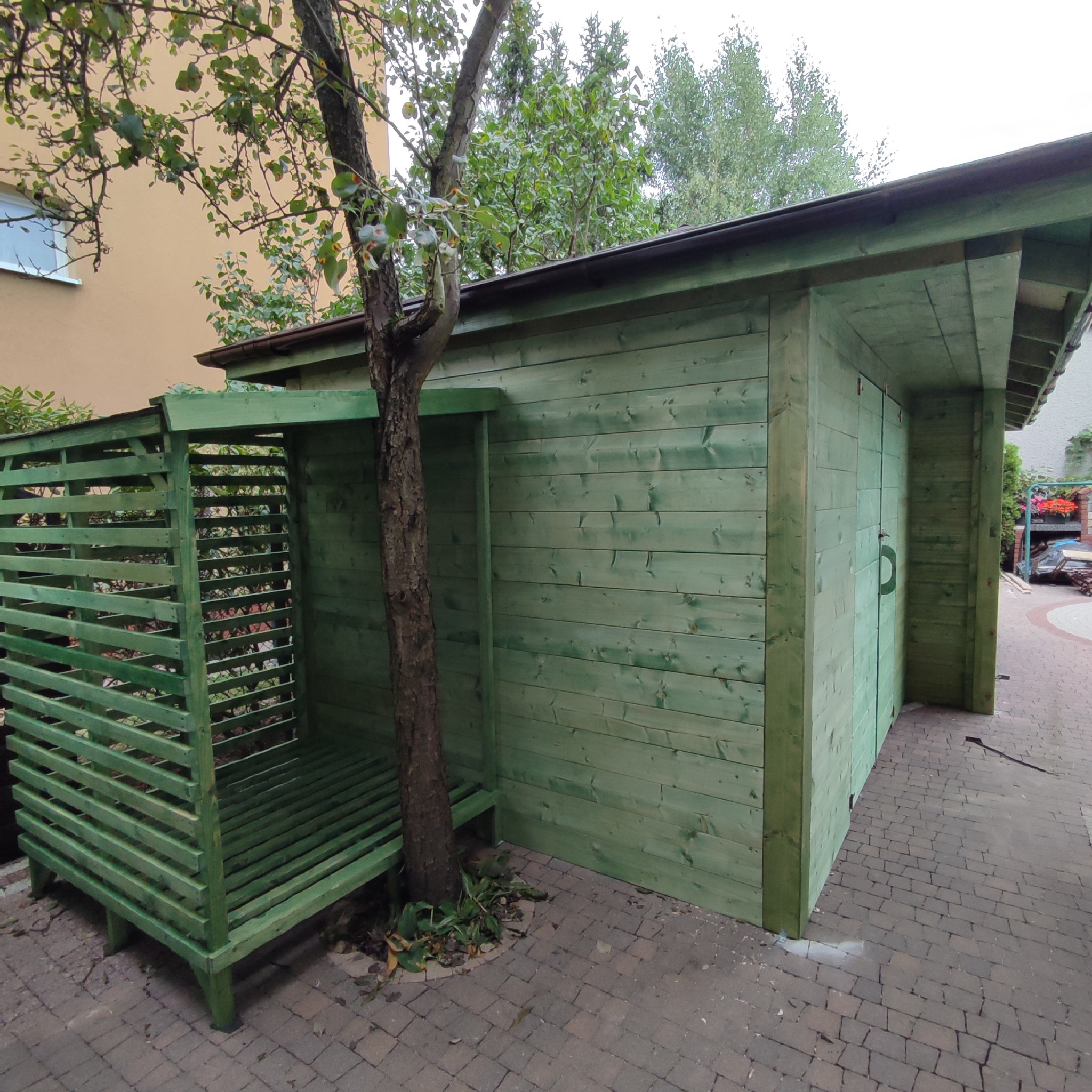 Zewnętrzna, drewniana konstrukcja pomalowana na zielono, składająca się z zamykanego pomieszczenia i przylegającej ażurowej wiaty, z drzewem rosnącym blisko ściany.