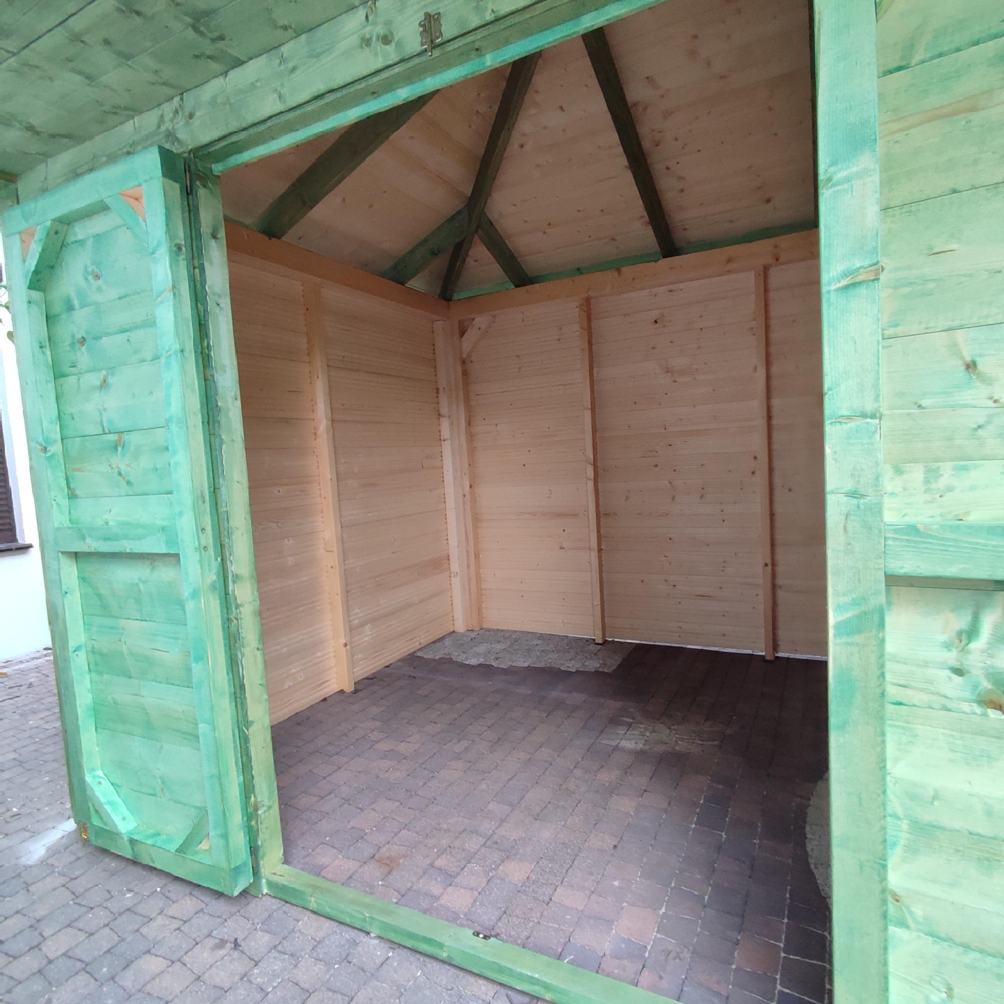 Wnętrze nowej, drewnianej konstrukcji z zielonymi elementami, ukazujące surowe ściany, podłogę z kostki brukowej i otwarte wejście.