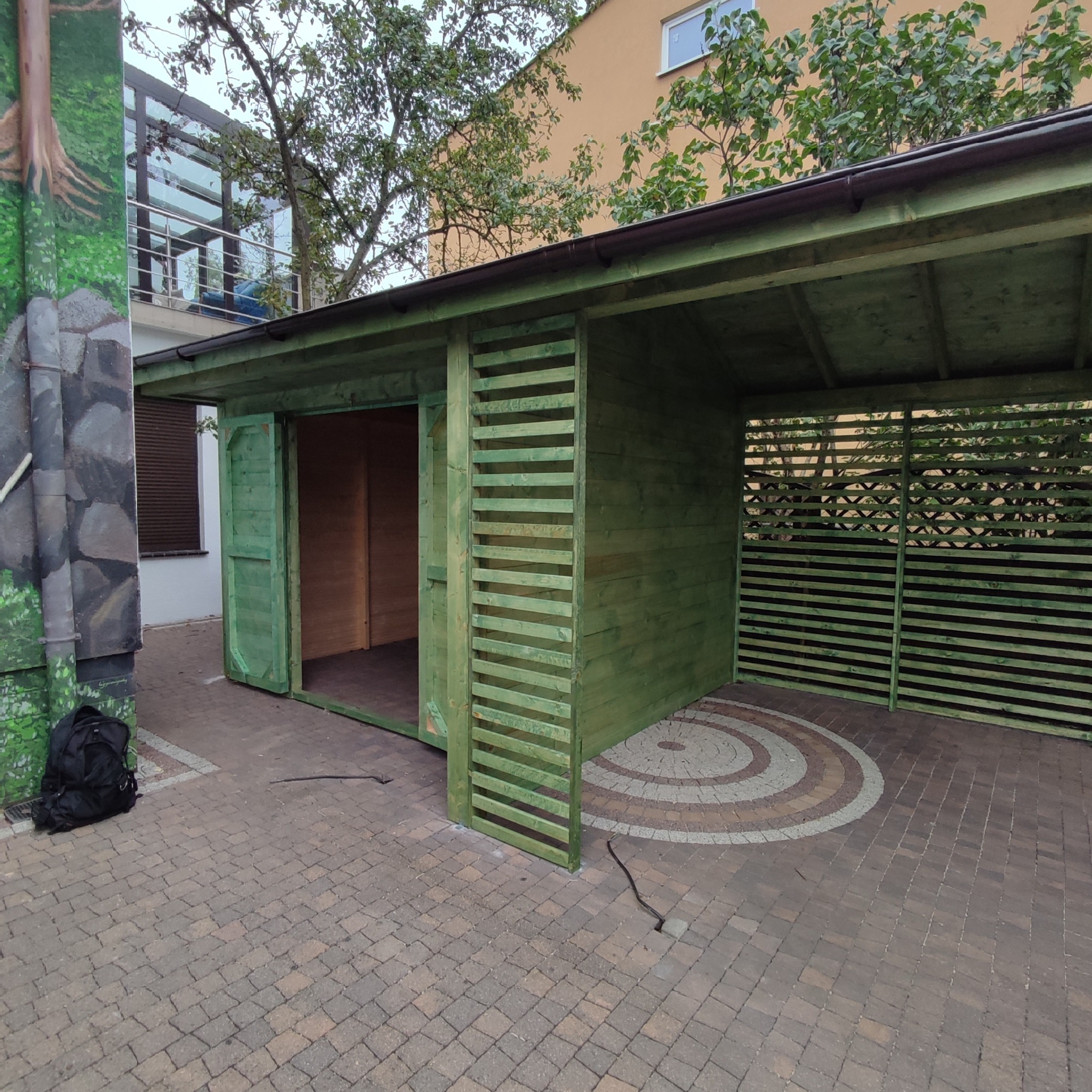 Zielona, drewniana konstrukcja z otwartymi drzwiami i ażurowymi ścianami na kostce brukowej z wzorem, obok ściana z muralem.
