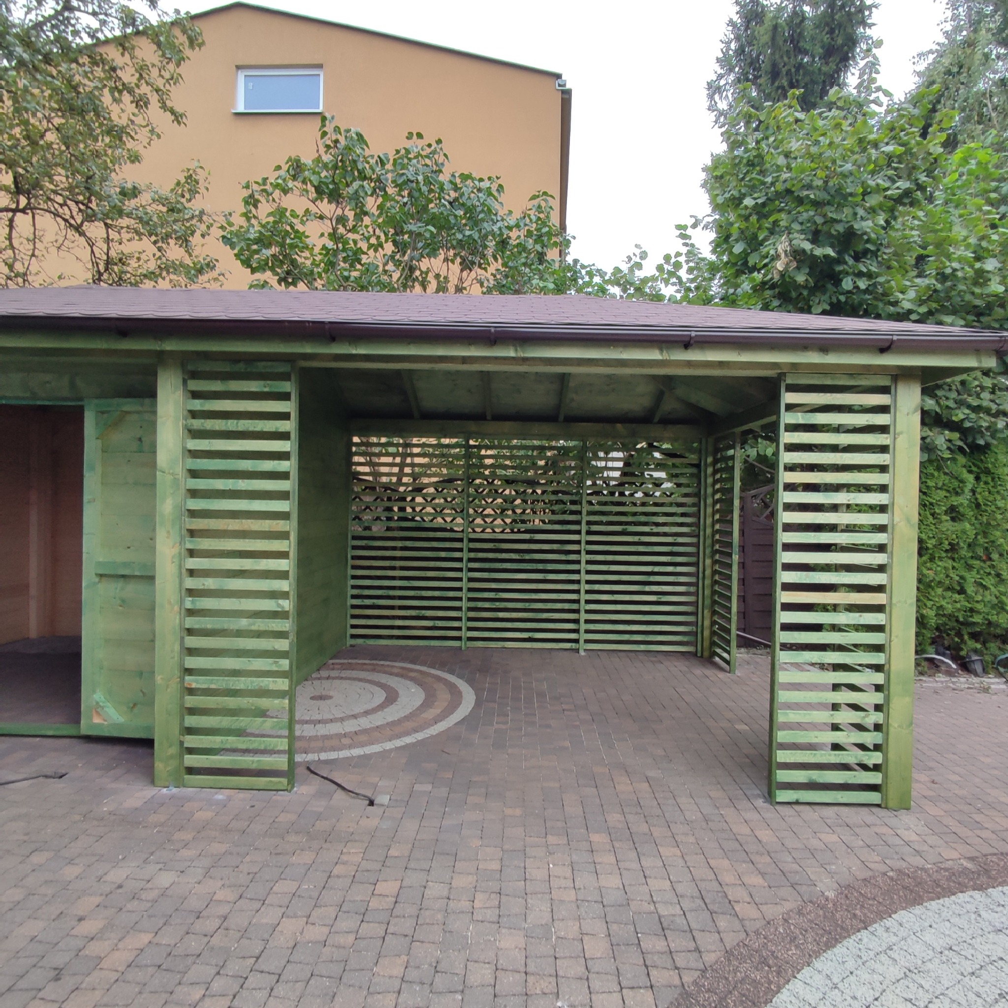 Zielona altana z drewnianymi listwami i brązowym dachem na kostce brukowej z ozdobnym wzorem, widok na budynek w tle.