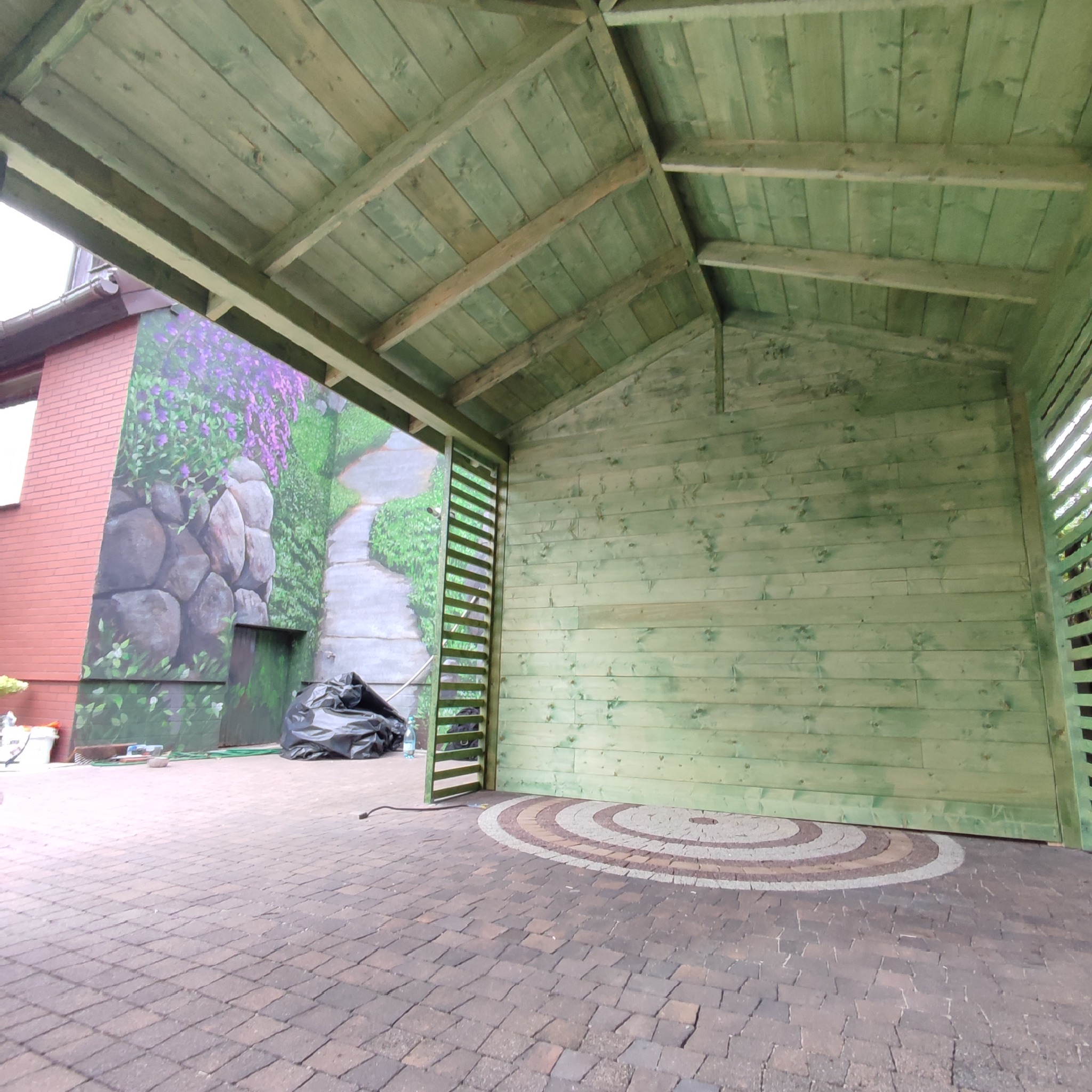 Drewniana konstrukcja z zielonymi deskami, widoczny fragment ściany budynku z malowidłem, na podłodze kostka brukowa z dekoracyjnym wzorem, w tle czarny worek na śmieci.