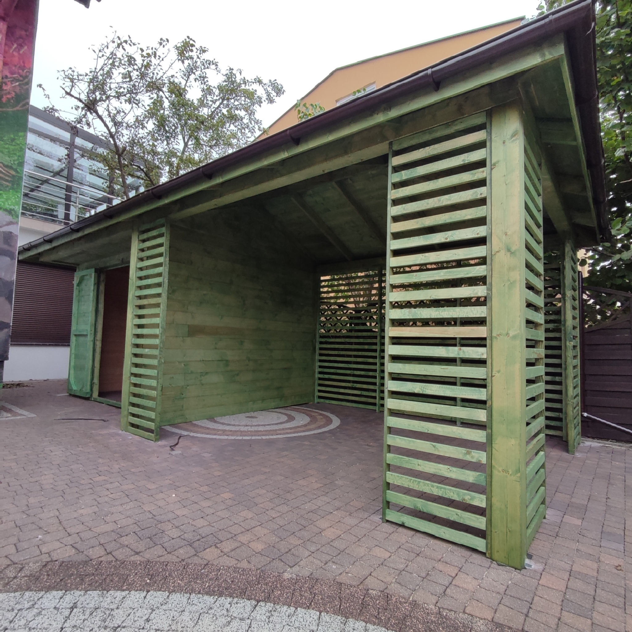 Drewniana konstrukcja z poziomymi listwami, pomalowana na zielono, z widocznym dachem i otwartą przestrzenią w środku, umieszczona na brukowanej powierzchni.