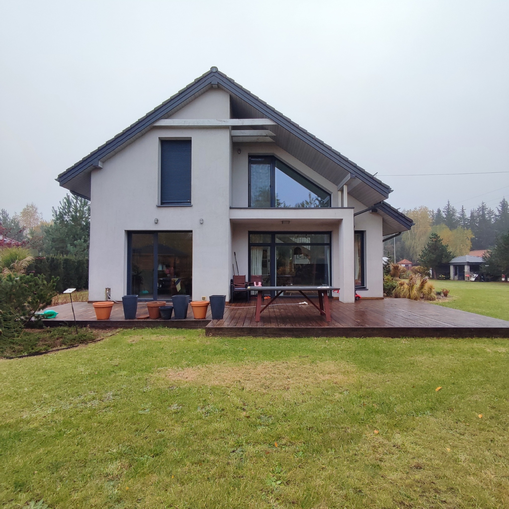 Nowoczesny dom z dużym drewnianym tarasem, na którym stoi stół i donice z kwiatami, widok z ogrodu w pochmurny dzień.