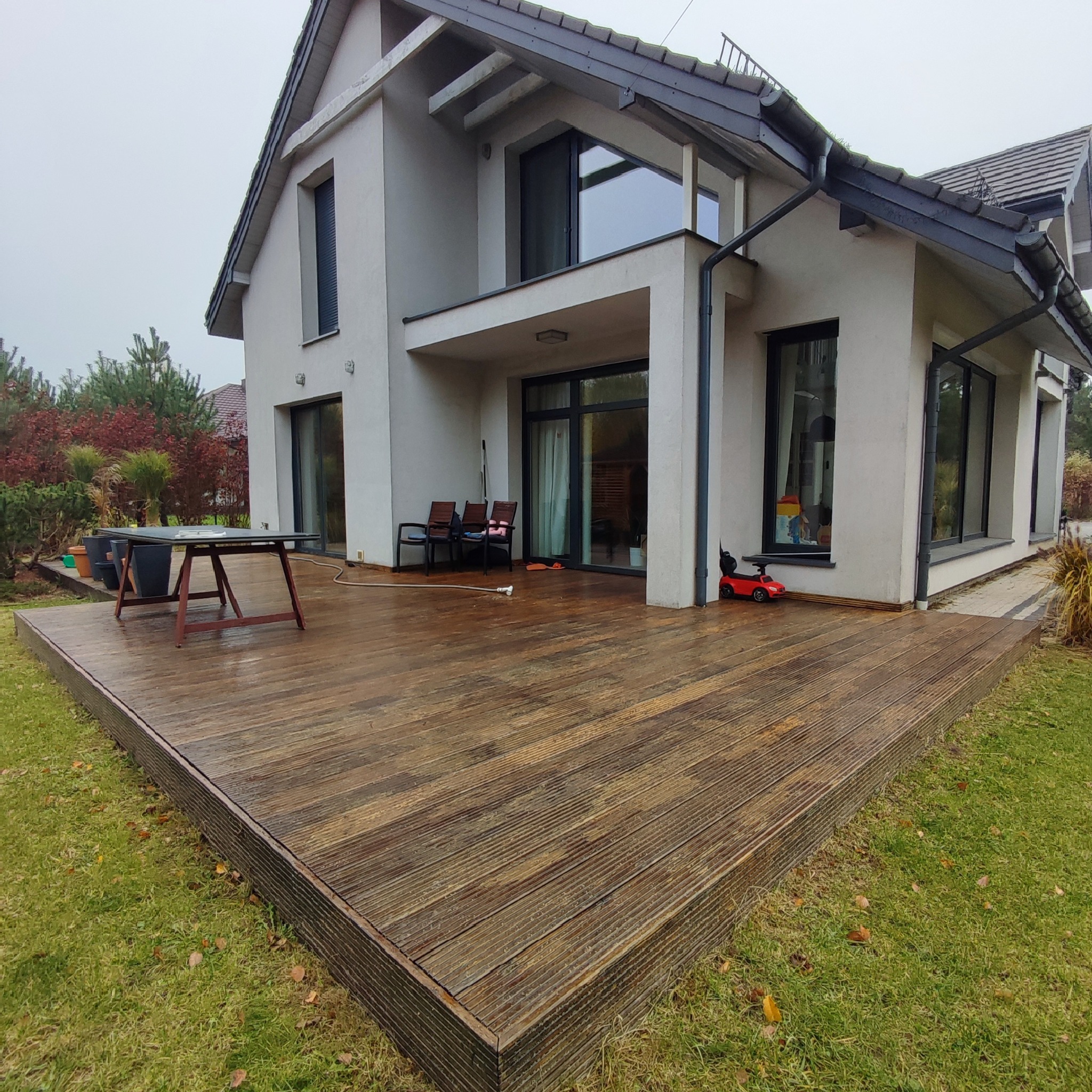 Rozległy taras drewniany przylegający do nowoczesnego domu z szarą elewacją, widoczny stół, krzesła i zabawka dla dziecka, otoczony zielonym trawnikiem i roślinnością w pochmurny dzień.