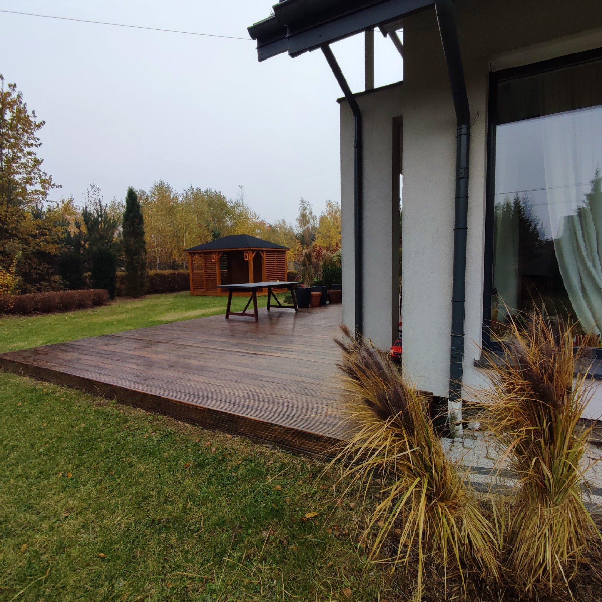 Zadaszony taras z drewnianą podłogą i stołem na zewnątrz nowoczesnego domu, z widoczną w tle altaną w ogrodzie.