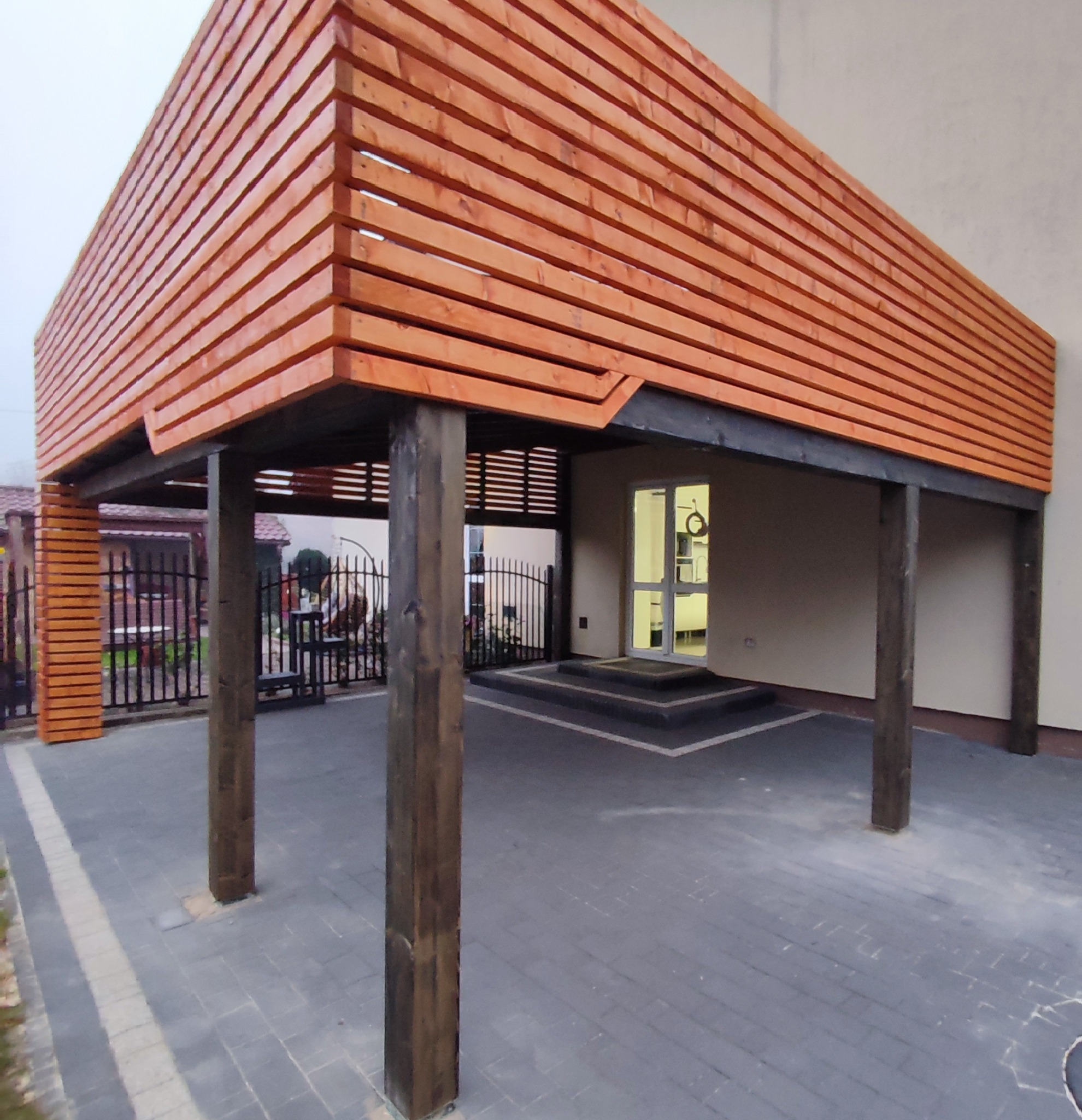 Zadaszenie tarasu wykonane z poziomych desek w kolorze jasnego drewna, wsparte na ciemnych, drewnianych słupach; widoczna kostka brukowa i wejście do budynku z oświetlonym wnętrzem.