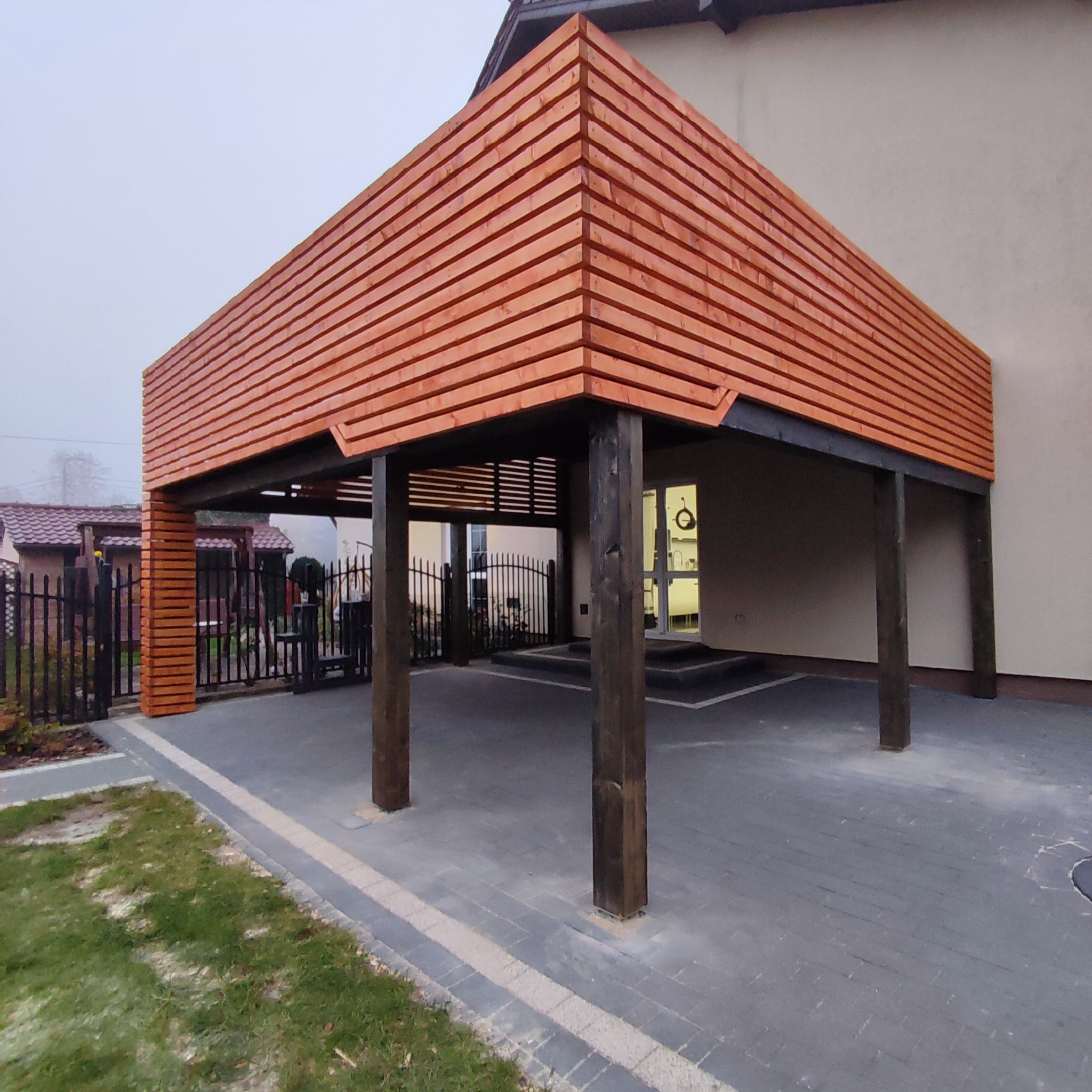 Zadaszenie tarasu wykonane z poziomo ułożonych desek w odcieniach brązu, wsparte na solidnych, ciemnych, drewnianych słupach. Widoczny fragment posesji z kostką brukową i trawnikiem.