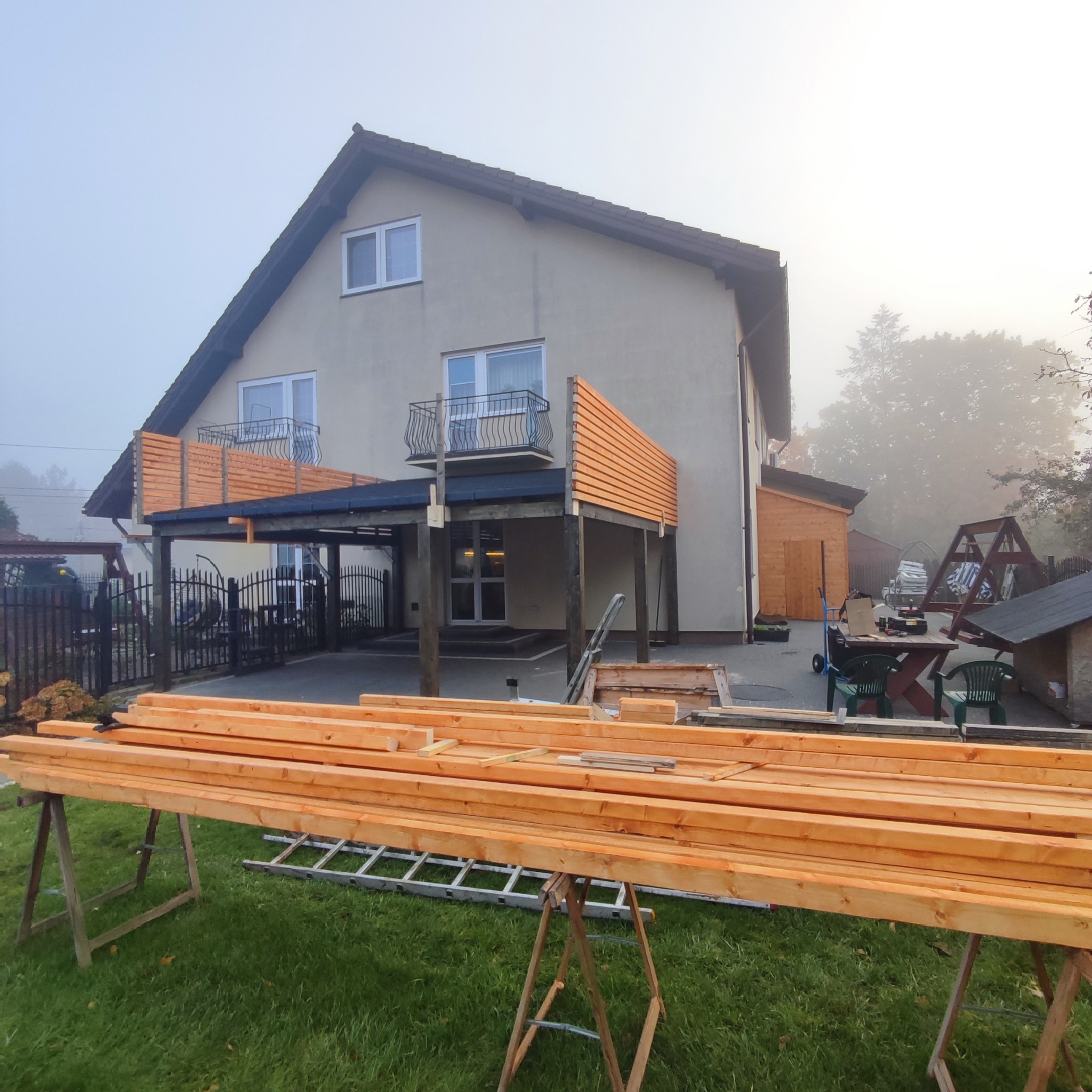 Budowa zadaszenia tarasu przy domu jednorodzinnym w porannej mgle; widoczne przygotowane deski, rusztowanie, częściowo ukończona konstrukcja z drewnianą balustradą.