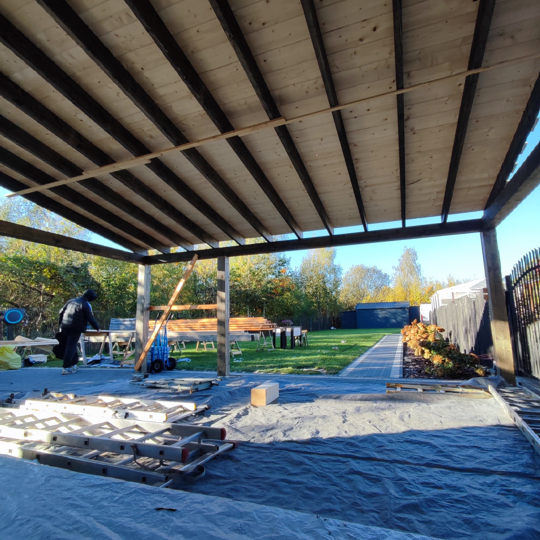 Montaż drewnianej konstrukcji z widocznym szkieletem dachu, drabiny, narzędzia i pracownik w tle, na zewnątrz w ogrodzie.