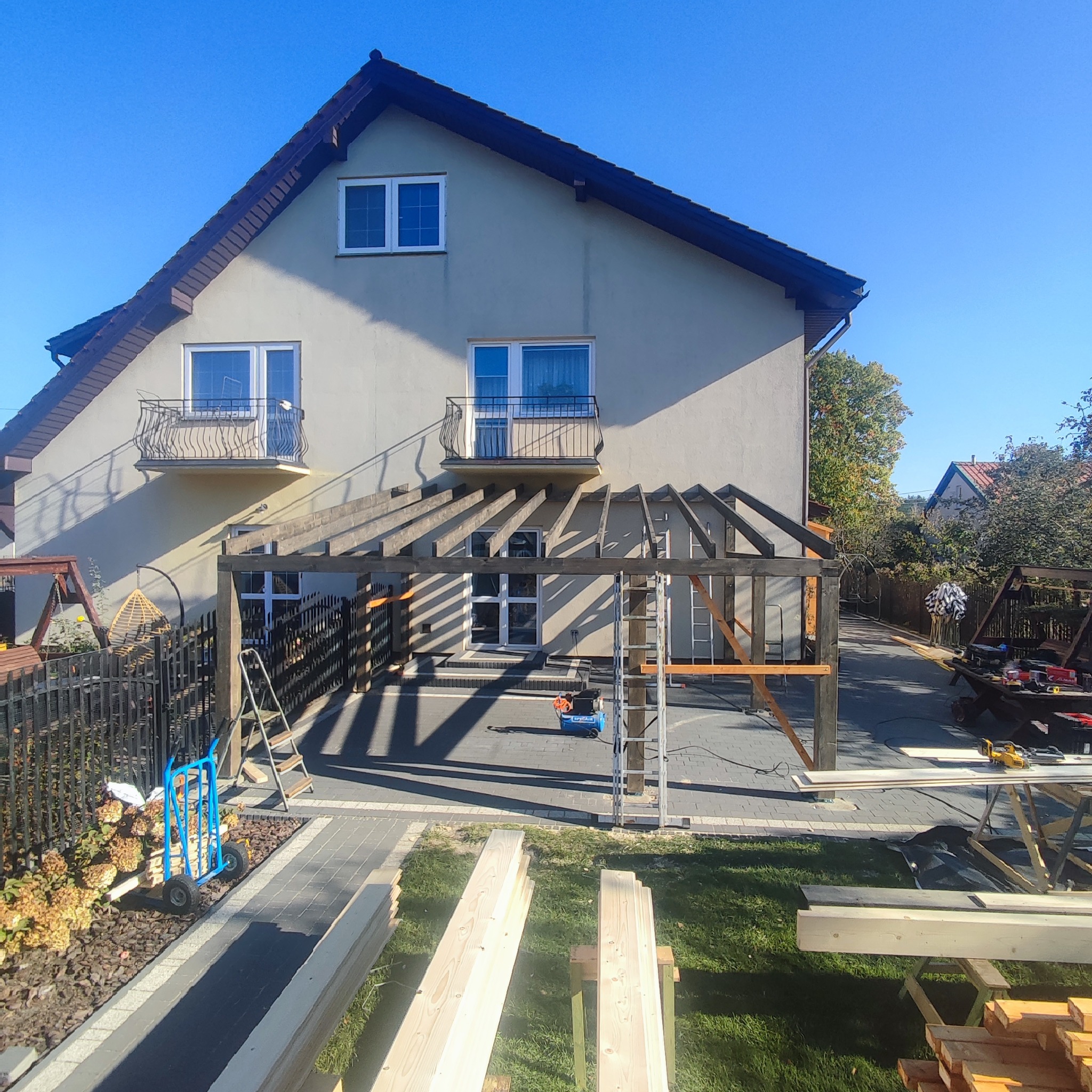 Konstrukcja drewniana zadaszenia tarasu w trakcie budowy przy domu jednorodzinnym, widoczne materiały budowlane, narzędzia i drabiny, jasne światło słoneczne.