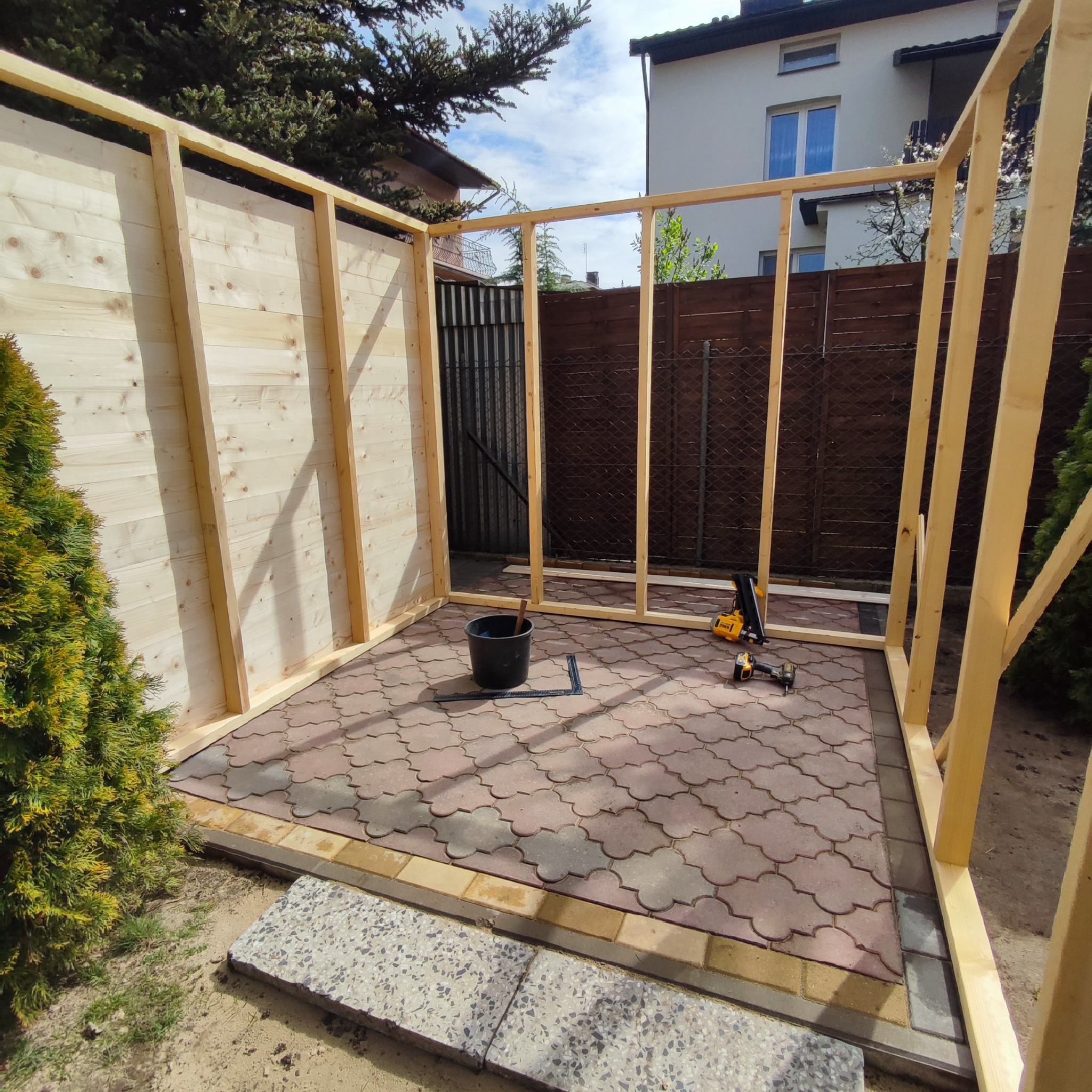 Konstrukcja szkieletowa z jasnego drewna, prawdopodobnie z desek, w trakcie budowy na utwardzonym podłożu z kostki, widoczne narzędzia i wiadro.