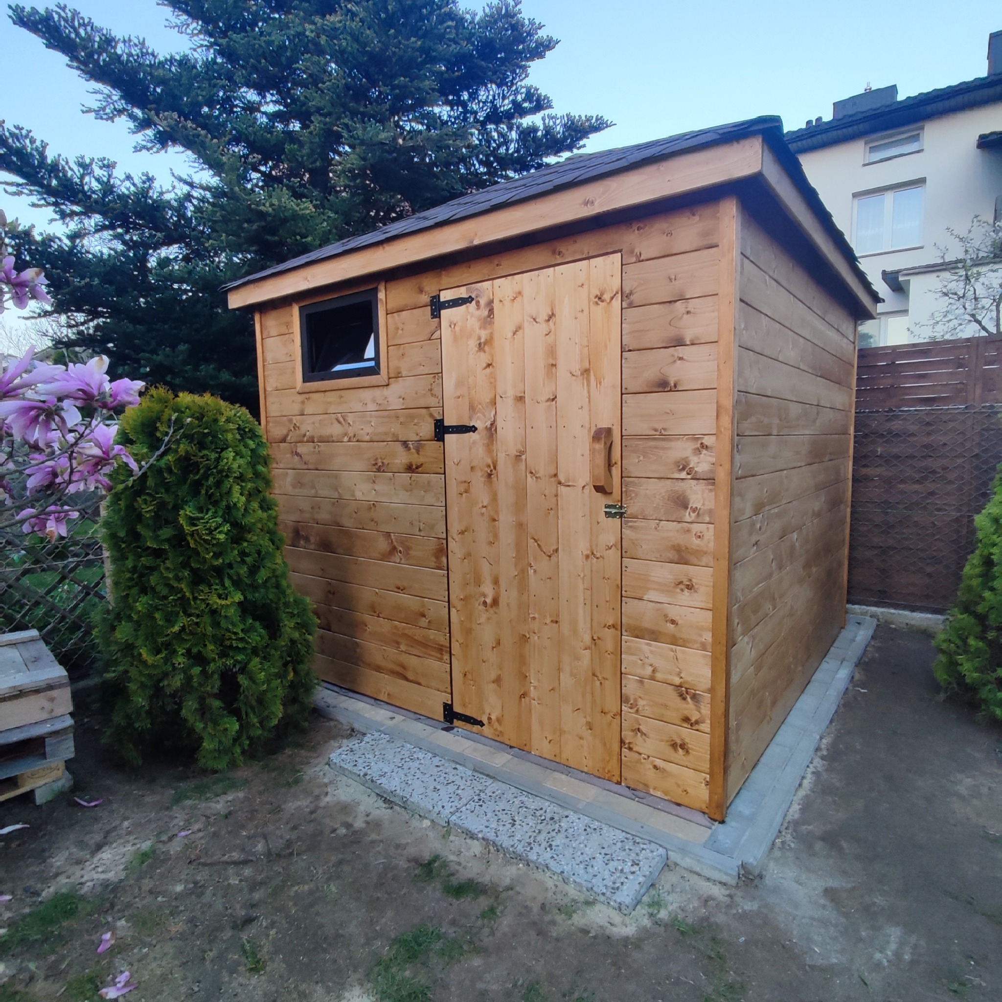 Drewniany domek narzędziowy z oknem i drzwiami na posesji, otoczony zielenią i kwitnącą magnolią.