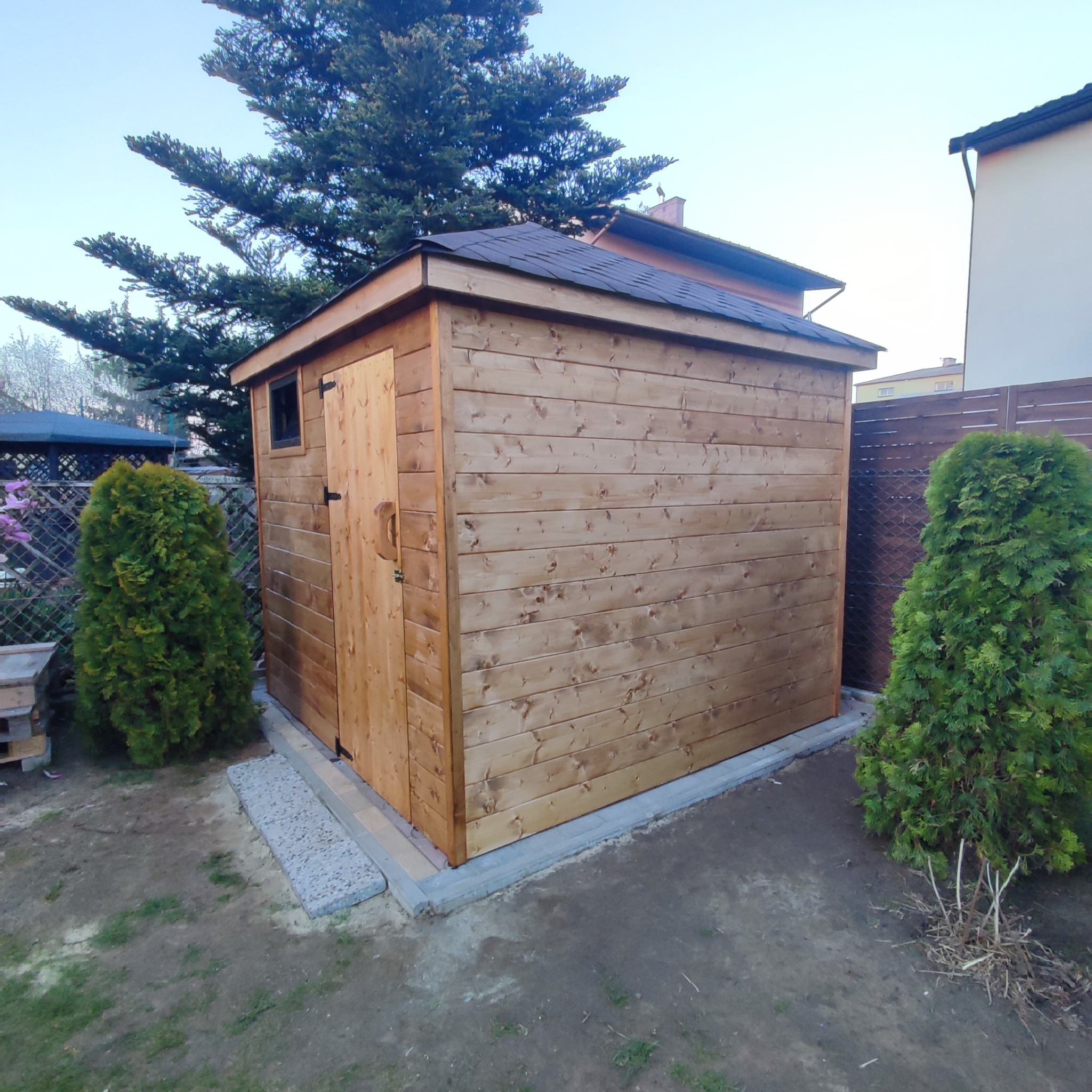 Drewniany domek narzędziowy z oknem i dachem z gontu bitumicznego, otoczony tujami i wykończony betonową podmurówką.