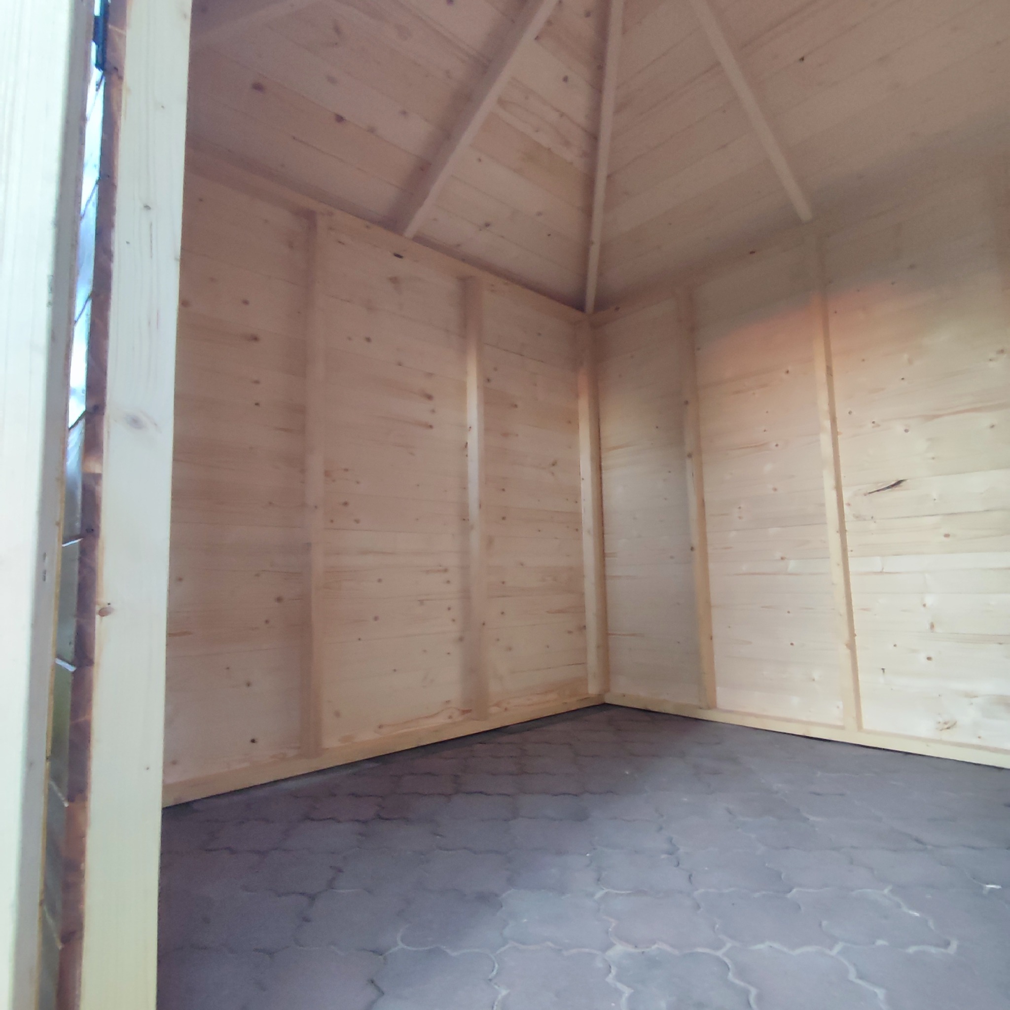 Wnętrze drewnianej konstrukcji z surowego drewna, widoczne ściany i dach, podłoga wyłożona szarą kostką brukową.