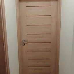 REKER Jarosław Ośliźlok - Jasne, drewniane drzwi wewnętrzne z poziomymi frezowaniami, zamontowane w korytarzu z drewnianą podłogą, widoczne z perspektywy podłogi.