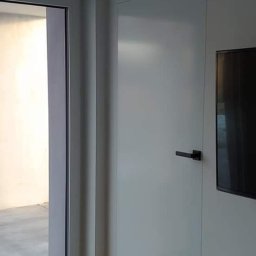 REKER Jarosław Ośliźlok - Nowoczesne, białe drzwi wewnętrzne z czarną klamką, zamontowane w minimalistycznym wnętrzu z drewnianą podłogą i widocznym fragmentem okna balkonowego.