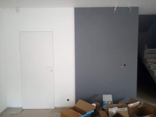 Świeżo zamontowane, białe drzwi wewnętrzne z ukrytą ościeżnicą, na ścianie pomalowanej na szaro widoczne puszki elektryczne, w tle schody i kartony po materiałach budowlanych.