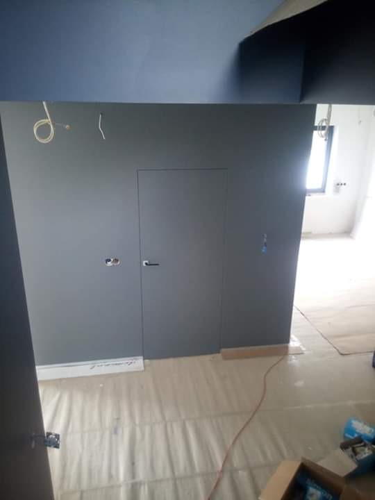 Szare ściany z ukrytymi drzwiami w trakcie remontu, podłoga zabezpieczona folią malarską, widoczne kable i puszki elektryczne.
