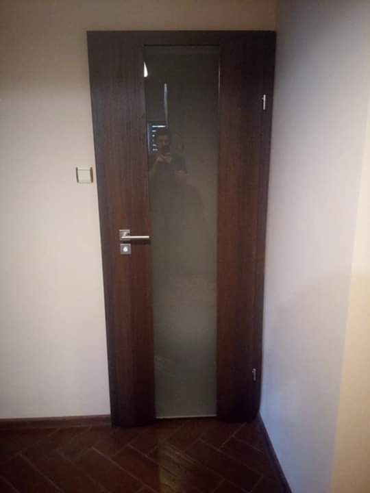Ciemne, drewniane drzwi wewnętrzne z matową szybą, widoczny numer '1' na górnej części ramy, odbicie fotografa w szybie.