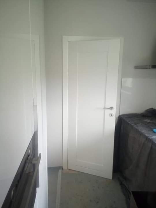 Nowo zainstalowane białe drzwi wewnętrzne z prostą klamką, widoczne w korytarzu obok zabudowy meblowej z szufladami, podłoga betonowa, zabezpieczone meble w tle.