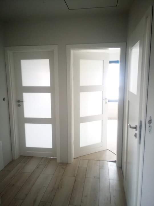 Dwa białe, przeszklone skrzydła drzwiowe z trzema panelami, osadzone w białych ościeżnicach, na podłodze z paneli imitujących drewno, jedno skrzydło otwarte, widok na jasne pomieszczenie w głębi.
