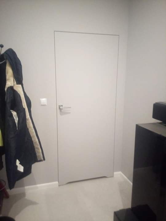 Białe, minimalistyczne drzwi ukryte w szarej ścianie, z widoczną klamką i kurtką wiszącą na wieszaku po lewej stronie.