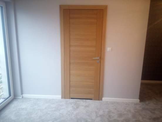 Nowo zamontowane, drewniane drzwi wewnętrzne z poziomymi panelami, srebrna klamka, widoczne na tle szarej ściany i jasnej podłogi.