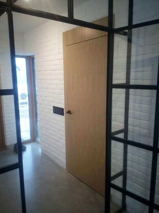 Minimalistyczna, gładka, drewniana wewnętrzna ościeżnica z czarną klamką na tle białej ściany z cegły, widoczna przez czarną, metalową ramę szklanej ściany działowej.