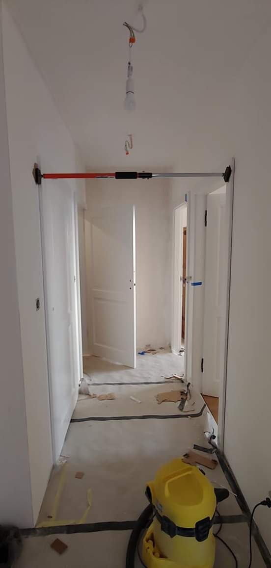 Korytarz w trakcie remontu, z zamontowaną rozpórką teleskopową do ościeżnic drzwiowych, widoczne przygotowane do montażu drzwi, zabezpieczona folią podłoga i odkurzacz przemysłowy.