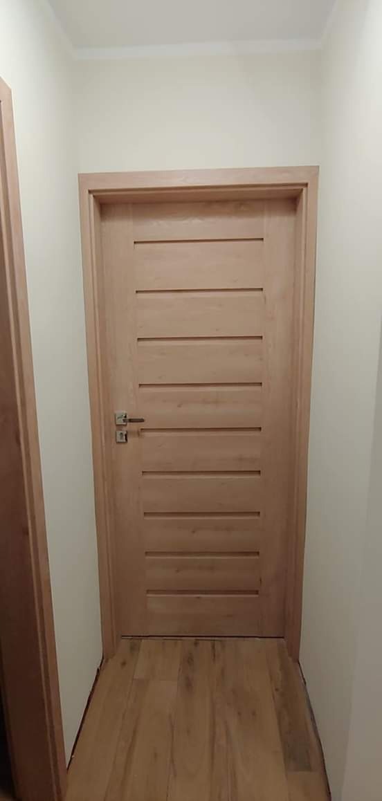 Jasne, drewniane drzwi wewnętrzne z poziomymi frezowaniami, zamontowane w korytarzu z drewnianą podłogą, widoczne z perspektywy podłogi.