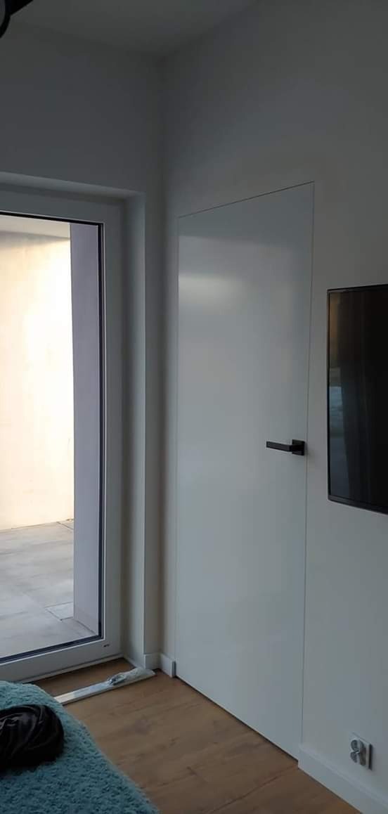 Nowoczesne, białe drzwi wewnętrzne z czarną klamką, zamontowane w minimalistycznym wnętrzu z drewnianą podłogą i widocznym fragmentem okna balkonowego.