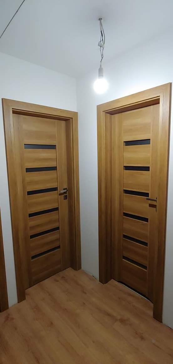 Dwa identyczne, drewniane drzwi wewnętrzne z poziomymi, czarnymi przeszkleniami zamontowane w jasnym pomieszczeniu z panelami podłogowymi, widoczna żarówka na kablu zwisająca z sufitu.