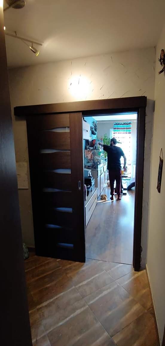 Zamontowane przesuwne drzwi z ciemnego drewna z poziomymi przeszkleniami, widok na wnętrze pokoju z osobą w tle i ułożoną podłogą z płytek imitujących drewno.