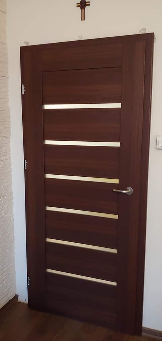 Ciemnobrązowe drzwi wewnętrzne z czterema poziomymi, jasnymi paskami, zamontowane w białej ścianie, widoczny krzyż na ścianie.