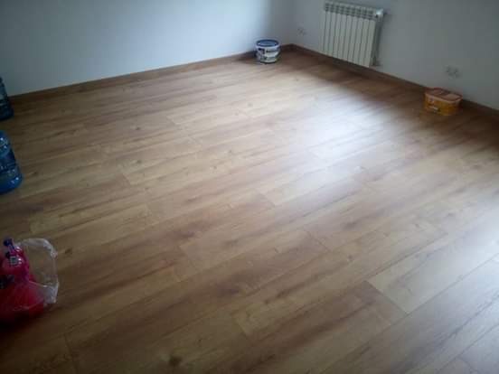 Nowo ułożona, jasnobrązowa podłoga z paneli laminowanych w pustym pokoju z grzejnikiem, wiadrami i butelkami z wodą w tle.